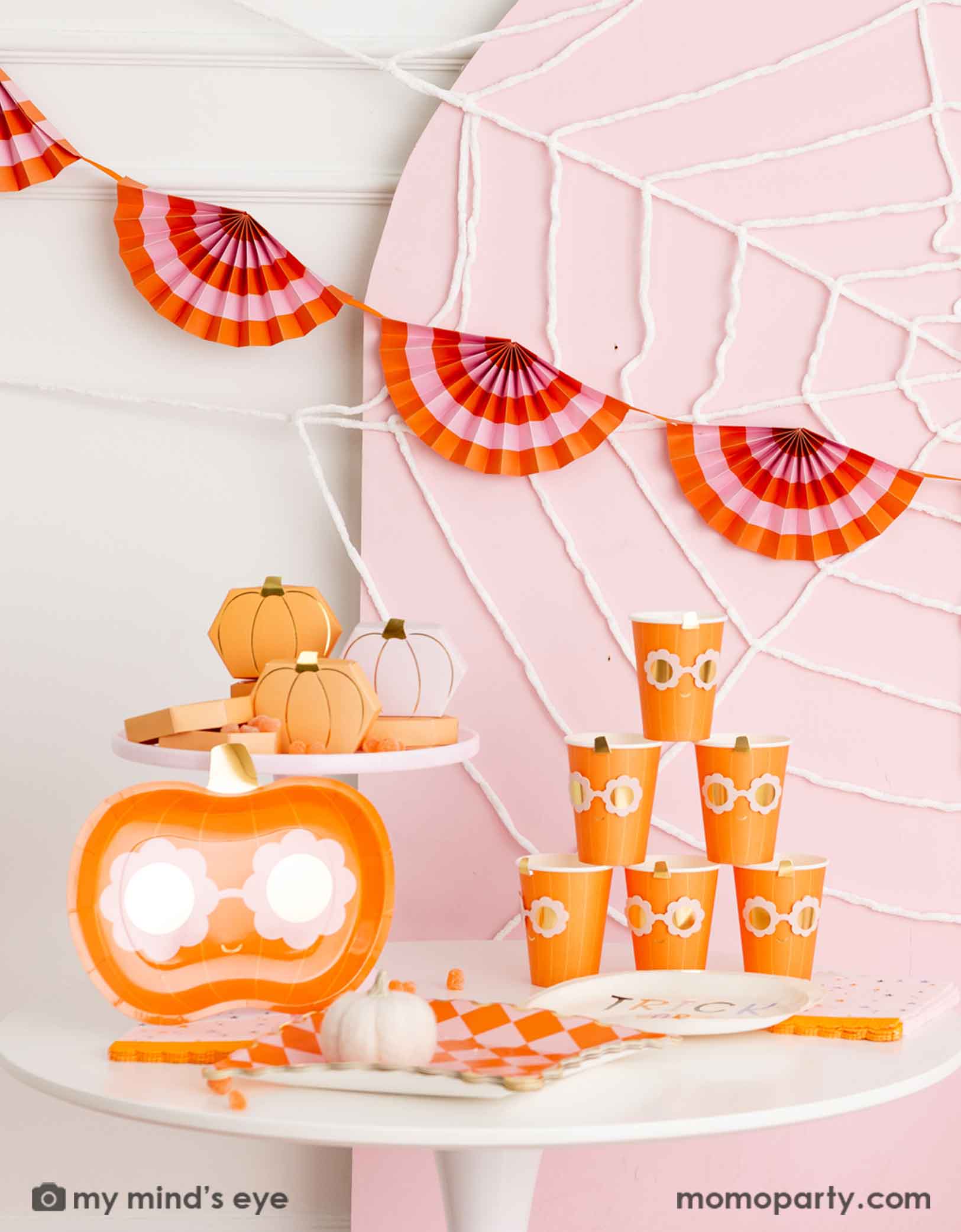A Halloween-themed party setup with a table filled with Momopartys's groovy pumpkin-shaped plates with daisy-shaped sunglasses, groovy pumpkin party cups, orange and pink checkered plates and pumpkin shaped treat boxes on a pink cake stand. In the back there's a pale pink wooden curved backdrop board decorated with spider web. A sweet kid's friendly Halloween themed decoration.  