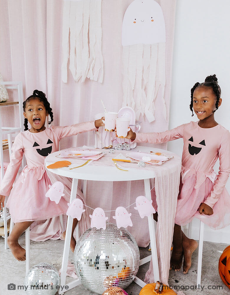 Two school aged girls in pink shirts that have Jack-o-lantern design on them and pink tutu sitting at a party table holding Momopartys's ghost shaped party cups. On the party table there are multiple pink Halloween party tableware spread out and the side is adorn with the pink ghost felt banner. Under the table there are some big disco balls and in the back hung some ghost paper decorations with streamer - a sweet and cute decoration for a Halloween celebration that is kid's friendly.
