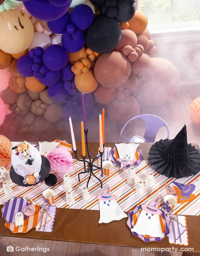 A festive pink Halloween party table set up featuring Momopartys's Bewitching ghost shaped plates, purple striped dinner plates, bewitching witch shaped hat napkins in three different colors of purple, orange and peach laid on a pink, orange and gold striped table runner. With some honeycomb pumpkins and witch hat decorations as the centerpiece, plus a whole of balloon garland in the colors of orange, brown, purple, making this an great inspo for a chic and cute Halloween party setup for this spooky season!