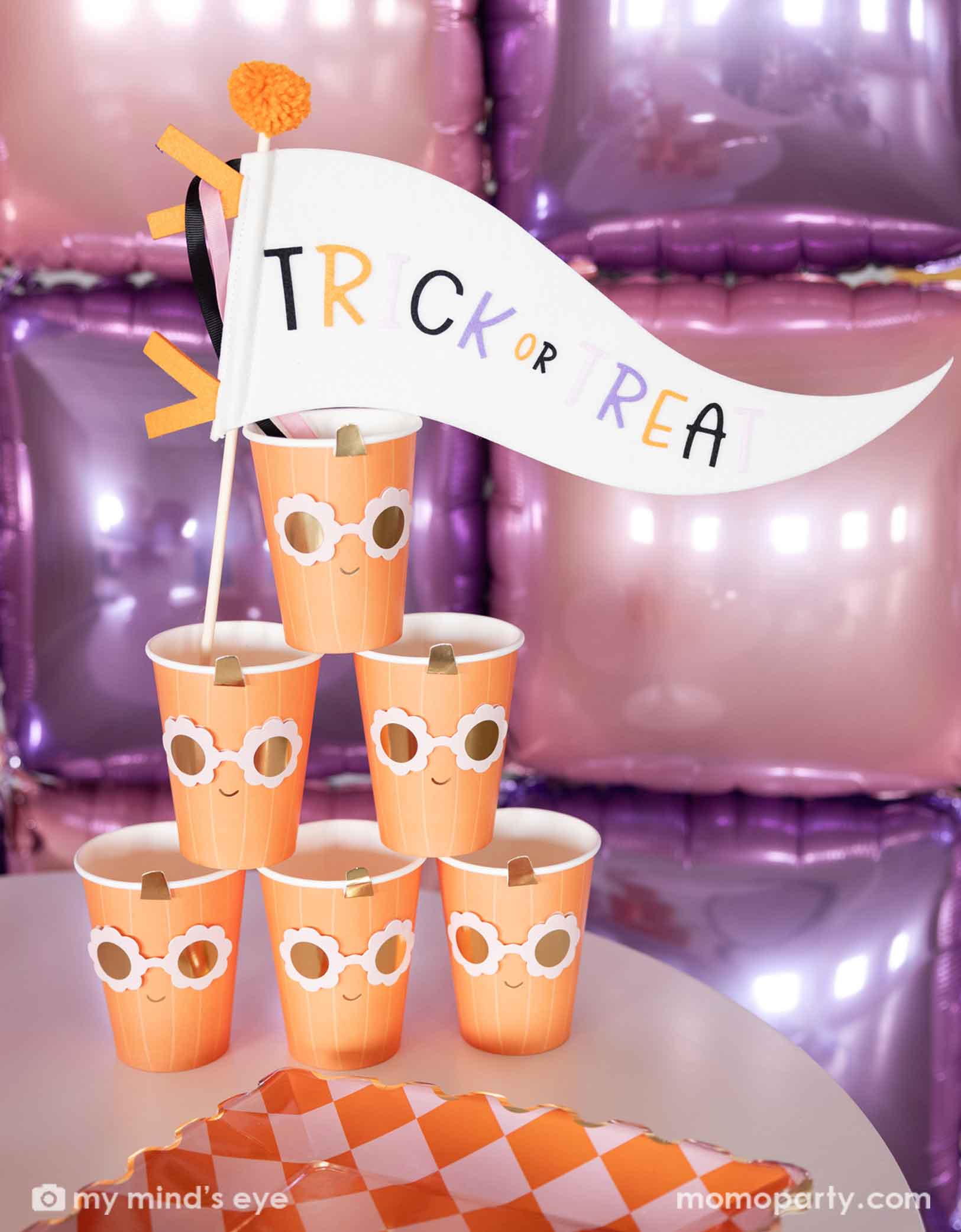 A stack of the orange pumpkin shaped paper party cups with daisy-shaped sunglasses from Momopartys with a 'Trick or Treat' felt party pennant on a Halloween inspired background with a foil balloon wall in pink and lilac. On the table there's  a orange and pink checkered dinner plate.