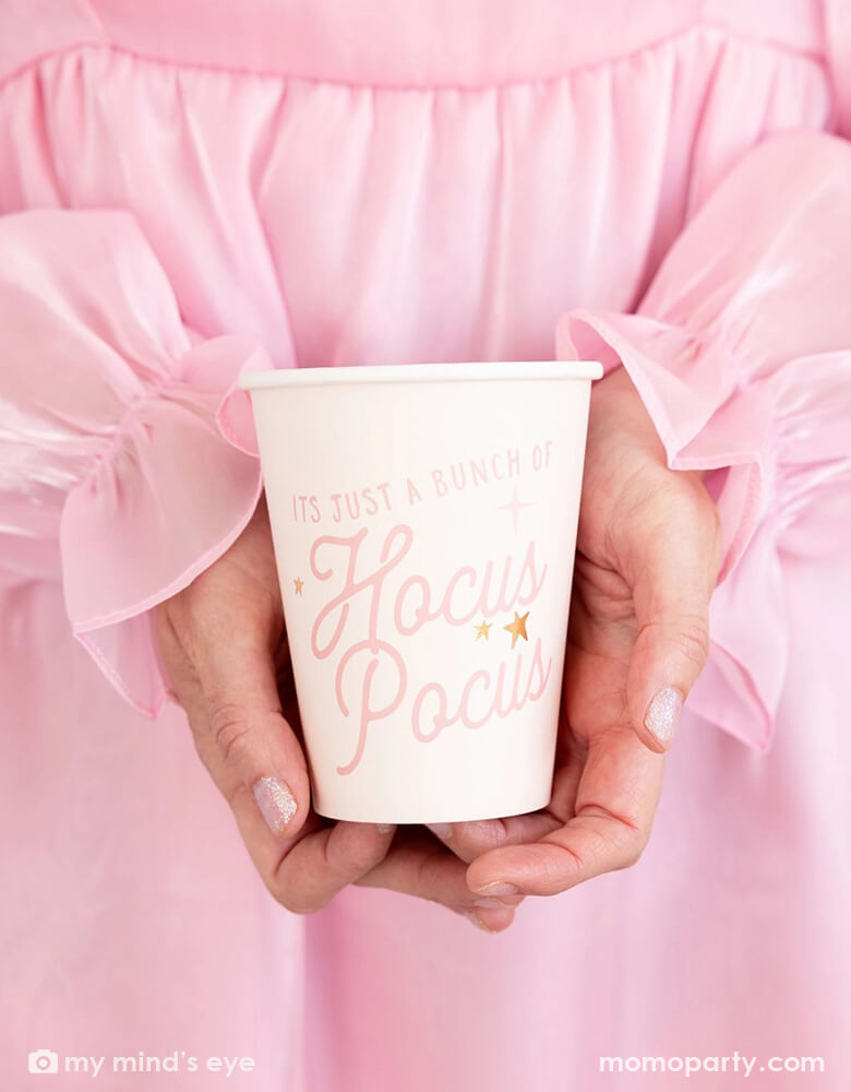 A lady in her pink dress holding Momopartys's 12 oz Hocus Pocus Pink Party Cup by My Mind's Eye. With "It's just a bunch of Hocus Pocus" written on the cup, and a playful pink design and a sprinkle of magic, this cup is perfect for any occasion. Spellbindingly stylish and practical, it's sure to be the life of your chic pink Halloween party in this spooky season!