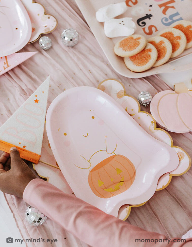 A Halloween party table filled with pink Halloween ghost themed party supplies from Momopartys including the pink ghost shaped paper plate with a jack-o-lantern and the 10" x 10" pink ghost scattered scalloped dinner plate. A little girl's hand in pink sleeve reaches out to the Boo felt party pennant. With mini disco ball decorations spread out and Halloween themed sugar cookies on the pink bamboo serving tray on the table, it makes a great inspo for a kid's friendly pink Halloween party this spooky season!