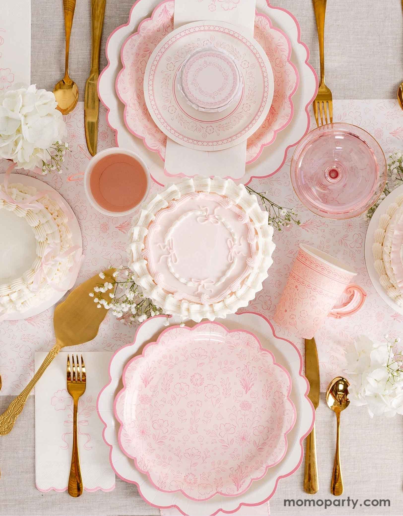An elegant tablescape featuring Momopartys's Pink Pembroke tableware collection including the 12" Pembroke Pink Edge Scallop Plates paired, 9" Pembroke Pink floral Plates, 7" Pembroke Pink Plates, Pembroke Pink napkins and party cups. Underneath the plates is the Pembroke Pink floral paper table runner. With the gold foil utensil set, a pretty floral arrangement and pastel pink butter cakes and glasses of rose champagne, this table setting is perfect for a spring Easter inspired celebration.