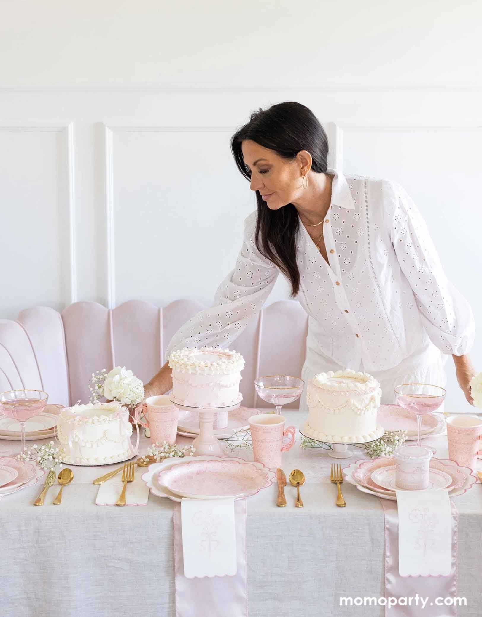 A lady is setting up an elegant party table featuring Momopartys's Pink Pembroke tableware collection including the 12" Pembroke Pink Edge Scallop Plate paired, 9" Pembroke Pink floral Plate, 7" Pembroke Pink Plate, the Pembroke Pink napkin and the matching party cup. With pretty floral arrangements, pink buttercream cakes and pink champagne glasses, this table setting is perfect for a spring inspired celebration, be it an Easter celebration, a bridal shower, a tea party birthday bash or Mother's Day party.