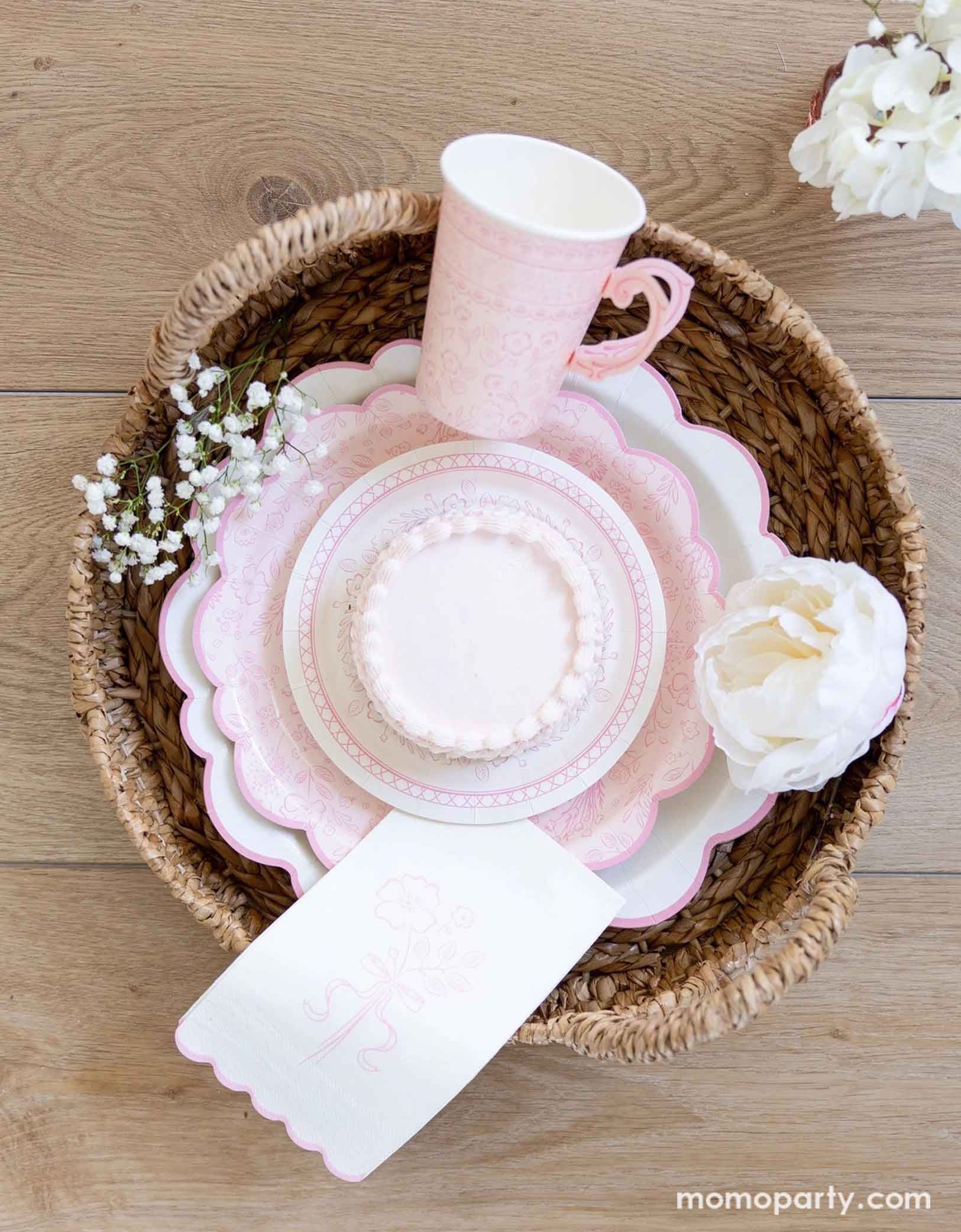 A woven basket filled with Momopartys's Pink Pembroke tableware collection including the 12" Pembroke Pink Edge Scallop Plate paired with 9" Pembroke Pink floral Plate and Pembroke Pink Edge Scallop Napkin. Underneath the plates is the Pembroke Pink floral paper table runner. With the gold foil utensil set, a pretty floral arrangement and pastel pink butter cakes and a glass of rose champagne,.