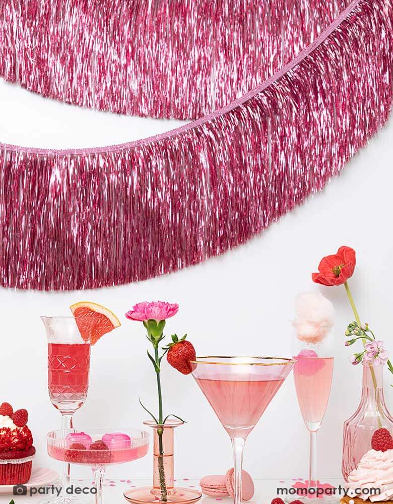 A pink themed party feature a sort of glasses with pink beverages and desserts including cupcakes, macaroons, candies and strawberries. The wall in back is decorated with Momopartys's 4.43 ft pink tinsel fringe garland by Party Deco. A perfect inspo for Valentine's Day or Galentine's Day celebrations.