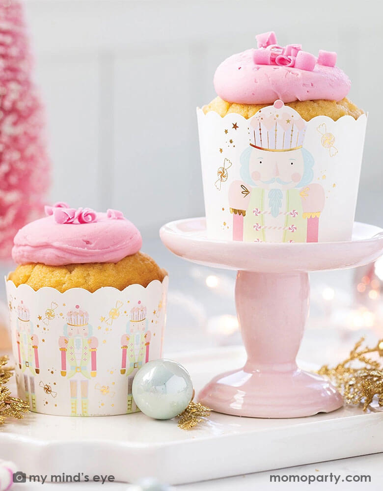 An adorable pink Christmas themed party table featuring pink cupcakes in Momopartys's gold Nutcracker food cups with gold foil accents on a small pink cupcake stand. Around the cupcakes there's some pink Christmas tree decorations in the back and some Christmas tree ornaments in gold and iridescent pastel colors. Making this a whimsical dreamy scene for a fabulous Holiday celebration.
