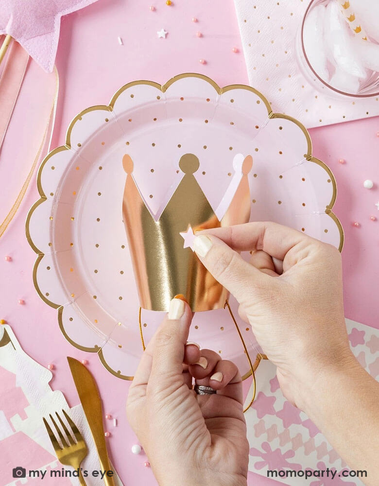 A girl's princess themed party table featuring Momopartys's pink princess themed party supplies including scalloped plates with gold polka dots, napkins, a pink star shaped wand and a gold paper crown being decorated with stickers, making this a great inspo for girl's princess themed birthday party celebration.