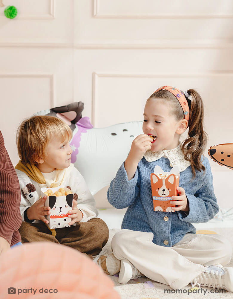 Two preschooler kids in a puppy dog themed birthday party sitting and holding Momopartys's puppy dog party snack boxes with adorable puppy design on them. They're eating the popcorn and chips from these cute puppy shaped treat boxes and having fun. In their back there are some cute dog shaped standing balloons.