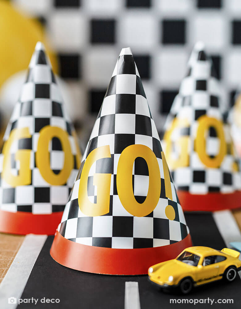 A close up shot of Momopartys's 4.3" x 6.3" race car checkered flag GO! party hats by Party Deco on party table decorated with a race car track sticker . Featuring the classic race car flag checkers with a red rim, each hat has a GO! written on it, making these a fun and festive addition to kid's race car themed celebration.