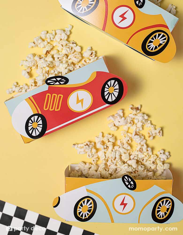 A festive bright yellow party table features three of Momopartys's race car snack boxes by Party Deco. These race car treat boxes are filled with popcorn, next to a race car flag checkered table runner, perfect for kid's car themed celebrations.