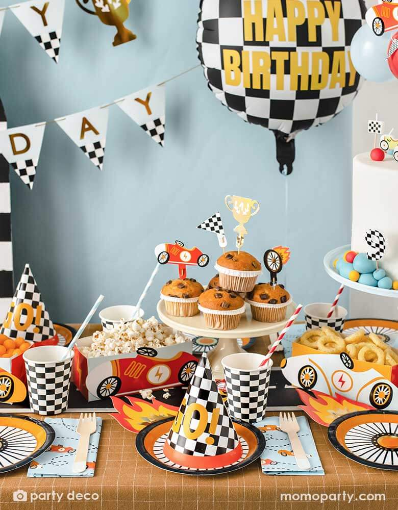 A kid's race car themed party table filled with race car themed party supplies from Momopartys including wheel shaped plates, checkered flag GO! party hats, blue race car large napkin, and checkered party cups. On the table there are muffins topped with race car themed cupcake toppers and vintage race car shaped snack holder filled with popcorn and onion ring chips. Above the table, there's a race car flag themed birthday banner and checkered happy birthday foil balloon.