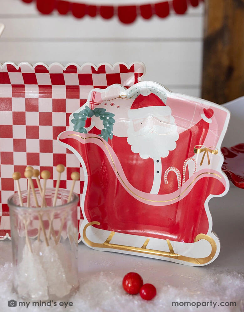 A table shot of Momopartys's 10" Pink and Red Checked Paper Plates next to the Santa on a sleigh shaped plate by My Mind's Eye, with some Holiday candies, gum balls and some artificial snow on the table as decoration. These playful Santa inspired plates add a touch of fun to any table setting. Get ready to serve up some serious style with these quirky and eye-catching plates this Holiday season!