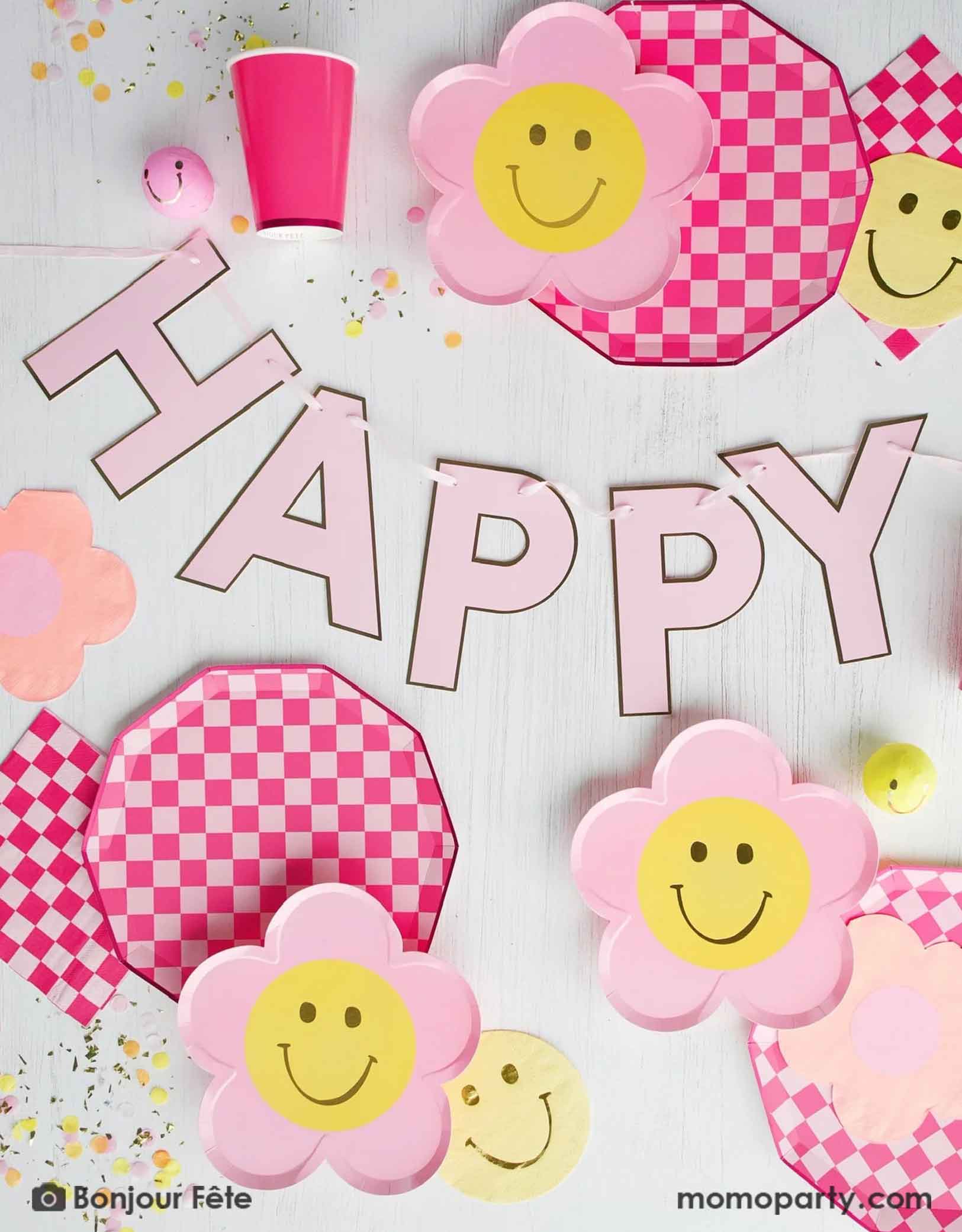 A retro inspired table setting for a kid's party featuring red and pink checkered paper plates with flower-shaped smiley faces, with a light pink "Happy Birthday" banner in the middle with confetti around from Momopartys on a light background.