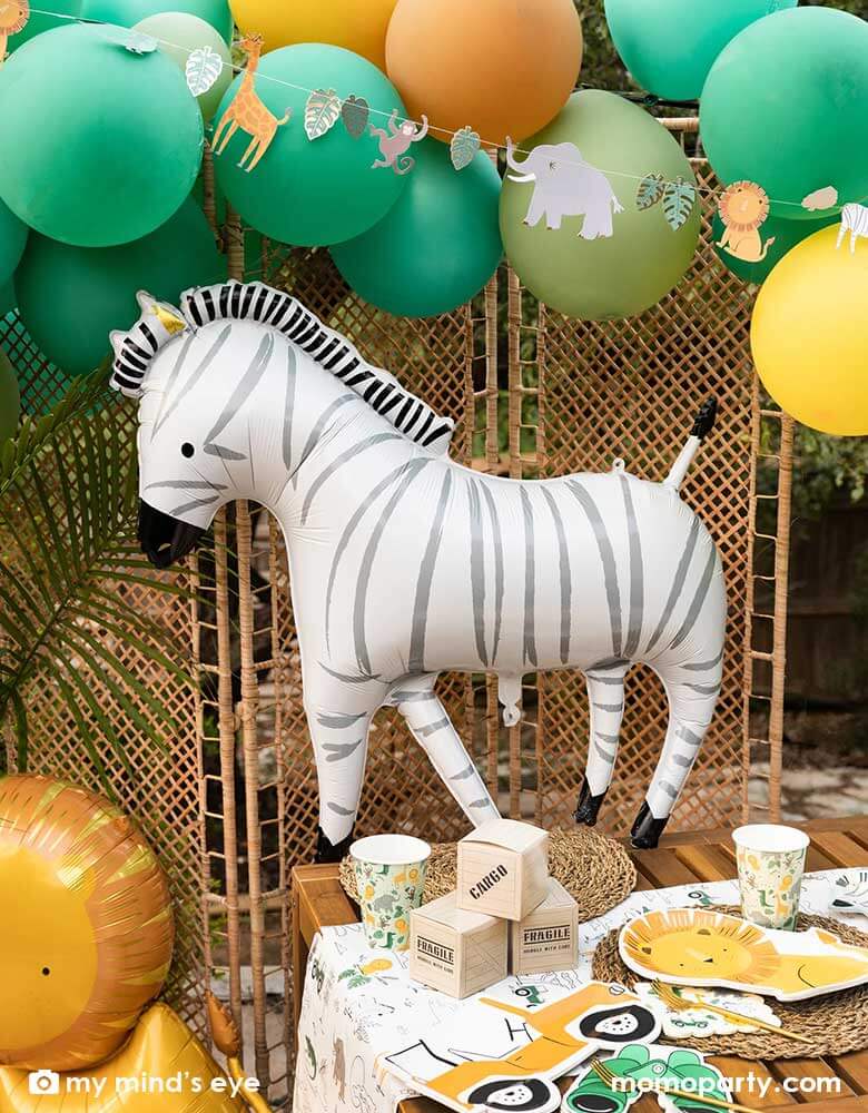 A festive kid's safari themed birthday party set up feature Momopartys's 24" zebra shaped foil mylar balloon by My Mind's Eye. Above it is a safari themed balloon garland in green, orange and yellow adorned with safari animal birthday banner. On the table, there are multiple safari themed tableware including a lion shaped plate, a Jeep car shaped plate, a binocular shaped dinner napkin and safari animal party cups on a safari themed paper table runner.