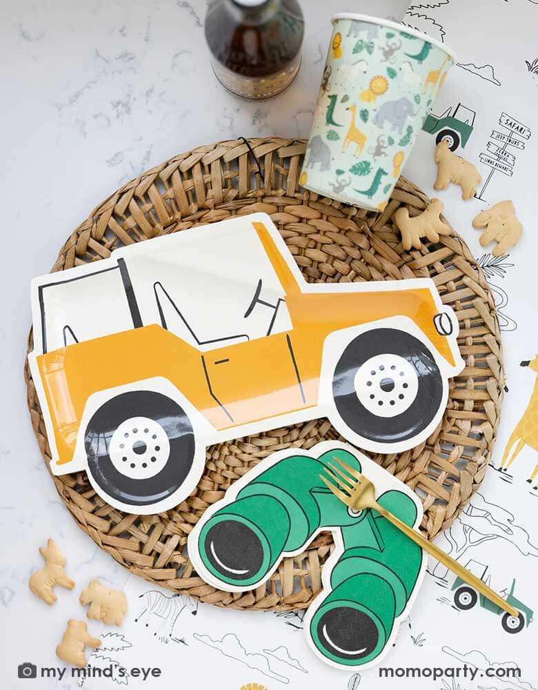 A safari adventure themed party table featuring Momopartys's mustard Jeep shaped dinner plate laid on top of a round bamboo weaved round tablemat which is placed on My Mind's Eye's safari adventure paper table runner featuring safari animals and safari landscape. Around the plate are a mint party cups with lots of safari animals and Momopartys's green binoculars shaped dinner napkins. With animal crackers on the table, makes this tablescape a great inspiration for kid's safari themed party.