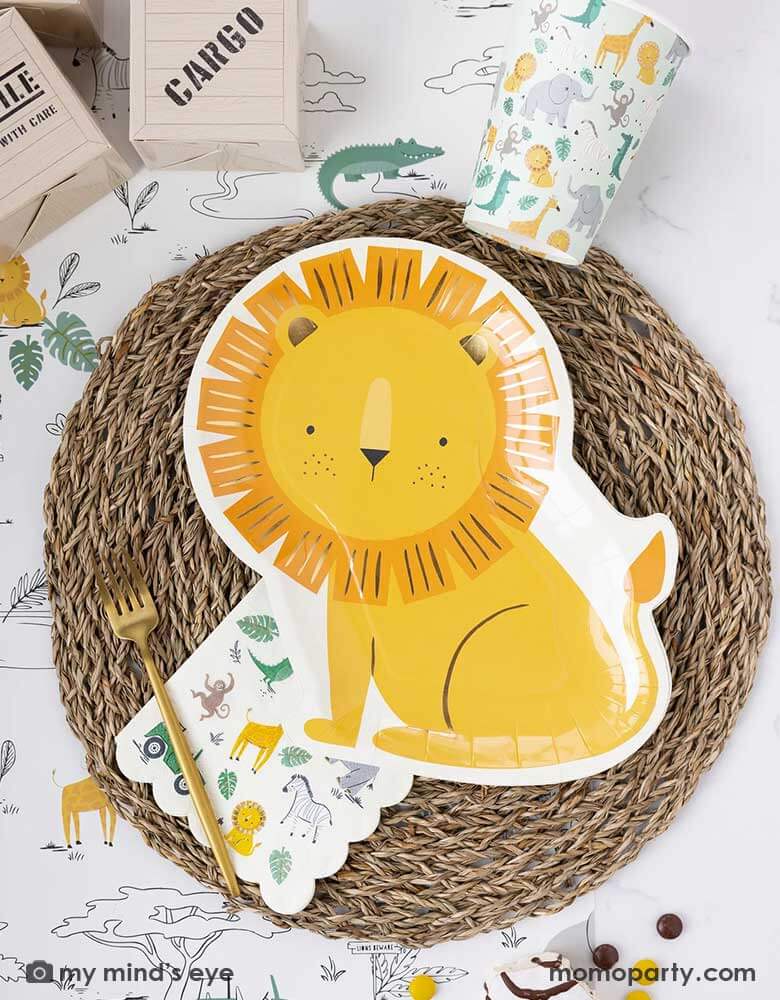 A safari adventure themed party table featuring Momopartys's 9" x 11" lion shaped plate laid on top of a round bamboo weaved round tablemat which is placed on My Mind's Eye's safari adventure paper table runner featuring safari animals and safari landscape. Around the plate are a mint party cups with lots of safari animals and the safari animal 5" x 5" small napkin. With My Mind's Eye's cargo favor boxes and zebra cakes on the table, makes this tablescape a great inspiration for kid's safari themed party.
