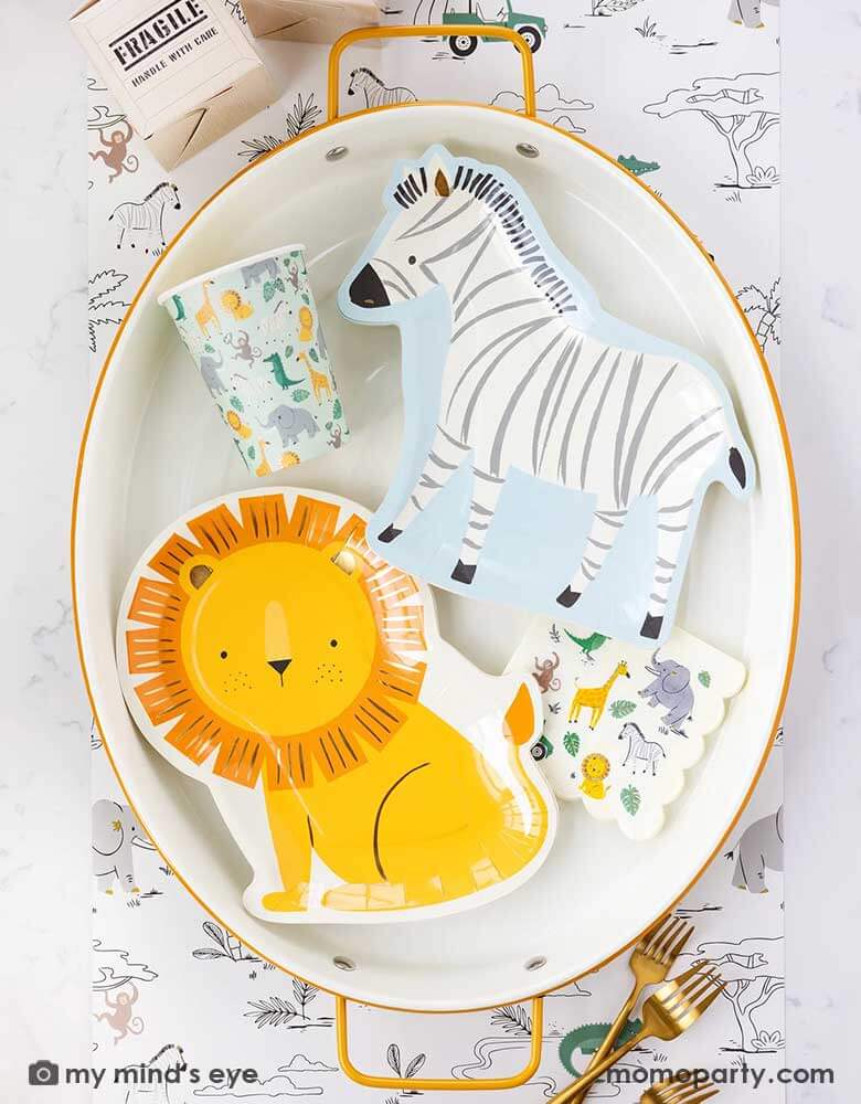 A safari adventure themed party table with an oval mustard edged plate with Momopartys's lion shaped plate and the zebra shaped plate, along with My Mind's Eye's safari party cups and small napkins, all laid on top of My Mind's Eye's safari adventure paper table runner featuring safari animals and safari landscape. Around the plate With My Mind's Eye's cargo favor boxes and gold utensils on the table, makes this tablescape a great inspiration for kid's safari themed party.
