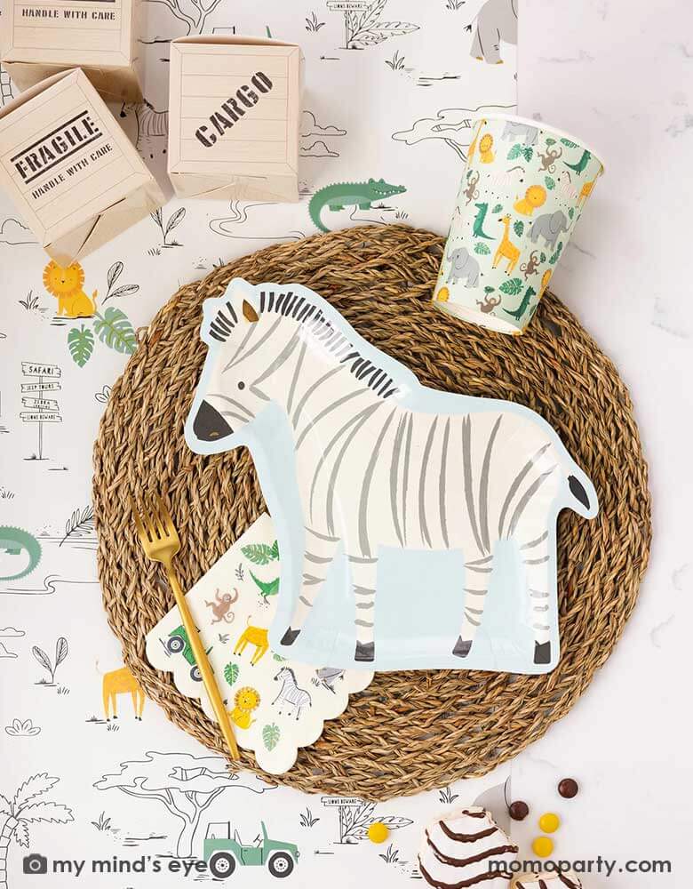 A safari adventure themed party table featuring Momopartys's zebra shaped plate laid on top of a round bamboo weaved round tablemat which is placed on My Mind's Eye's safari adventure paper table runner featuring safari animals and safari landscape. Around the plate are a mint party cups with lots of safari animals and the safari animal 5" x 5" small napkin. With My Mind's Eye's cargo favor boxes and zebra cakes on the table, makes this tablescape a great inspiration for kid's safari themed party.