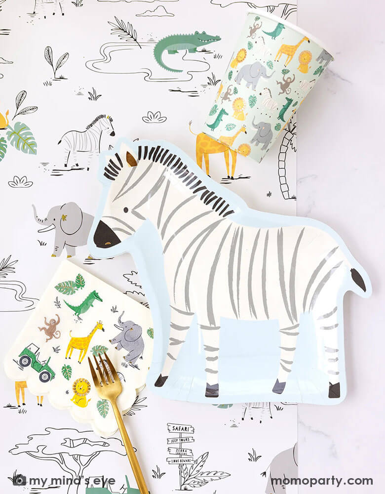 A safari adventure themed party table featuring Momopartys's zebra shaped plate by My Mind's Eye's and mint safari party cups and small napkins, all laid on top of My Mind's Eye's safari adventure paper table runner featuring safari animals and safari landscape. Around the plate With My Mind's Eye's cargo favor boxes and gold utensils on the table, makes this tablescape a great inspiration for kid's safari themed party.