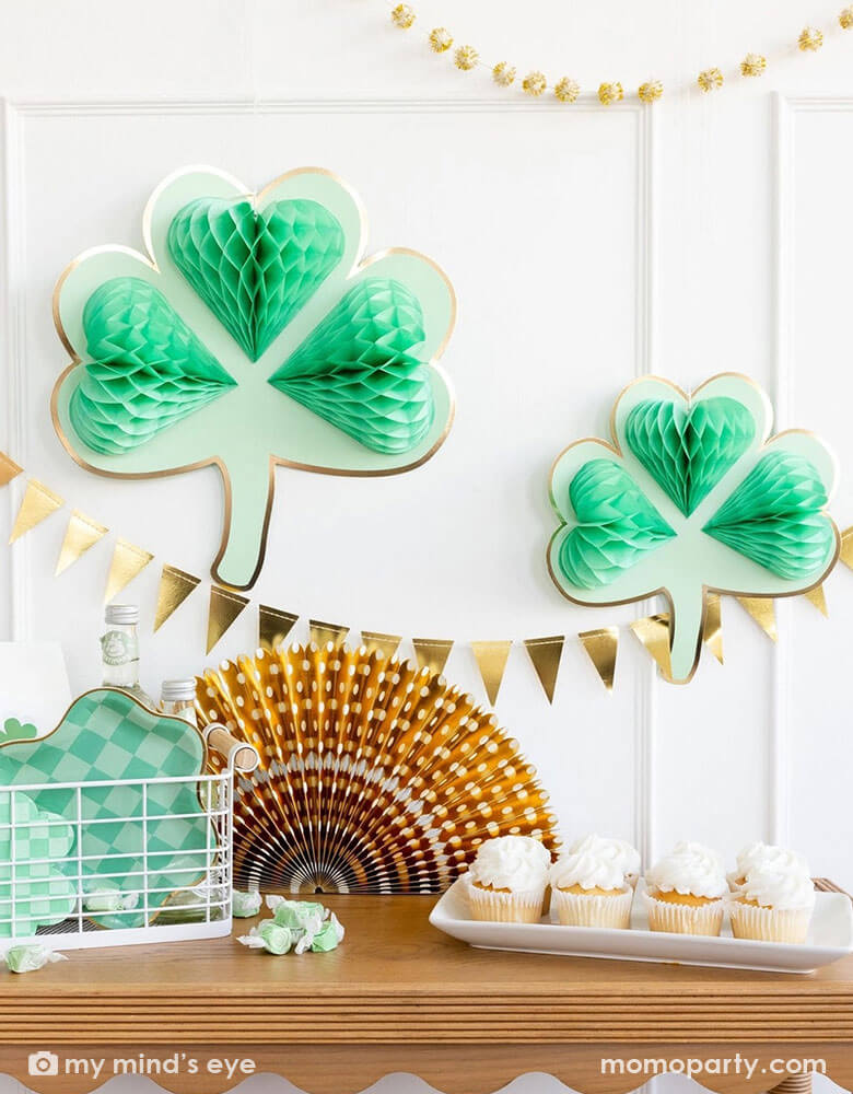 A corner of a room decorated with two of Momopartys's shamrock honeycomb decorations and a gold foil flag banner hung on the wall. On the cabinet counter, there's a gold foil paper party fan, a few cupcakes and a basket filled with Momopartys's St. Patrick's Day party tableware including scalloped paper plates, lucky paper plate set, checkered shamrock shaped napkins and plates next to some green taffy candies, makes this a perfect info for a festive St. Patrick's Day celebration.