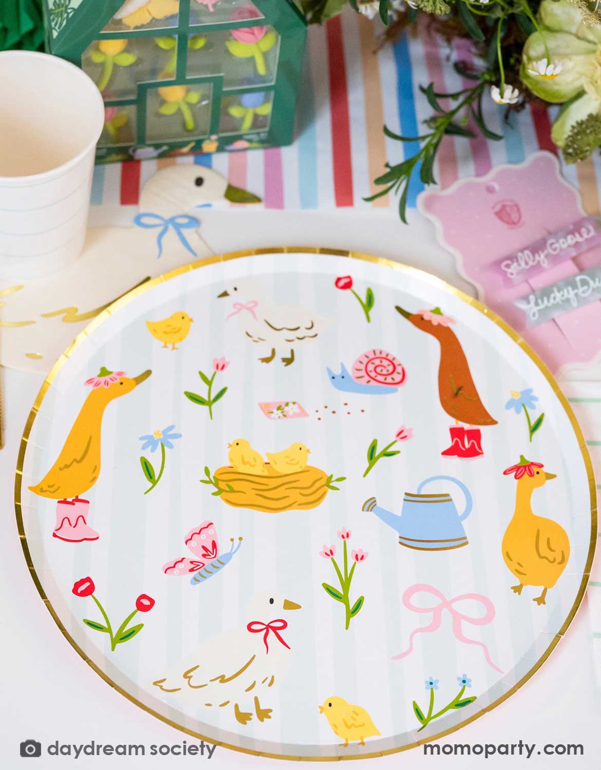 A closeup of Momopartys's 10" silly goose paper plate featuring adorable spring little animal illustrations in an Easter table setting with colorful flowers, tulip honeycomb decorations, goose themed party plates, napkins, party cups, pink cutlery and multiple kid's Easter small gifts with bunny designs on a colorful striped runner.
