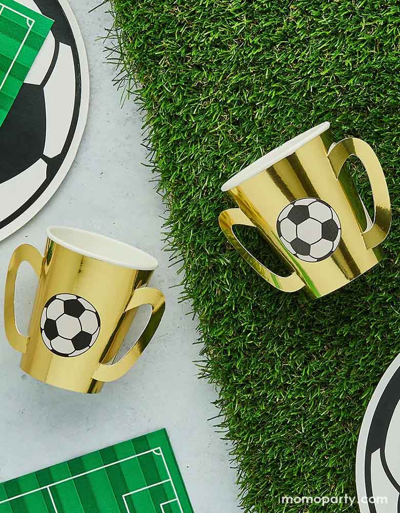 A soccer theme party table featuring soccer themed party supplies from Momopartys including gold trophy party cups with soccer ball design on them on a faux grass runner, surrounded by soccer ball shaped plates, soccer field shaped napkins, a perfect inspo for soccer themed gathering, including kid's soccer birthday parties and viewing parties.