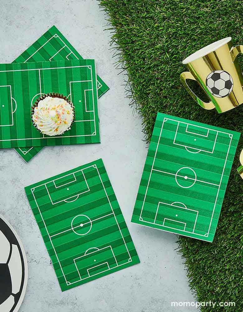 A soccer theme party table featuring soccer themed party supplies from Momopartys including soccer field dinner napkins on a faux grass table runner, surrounded by soccer ball shaped plates and gold trophy shaped party cups with soccer ball design on them, a perfect inspo for soccer themed gathering, including kid's soccer birthday parties and viewing parties.
