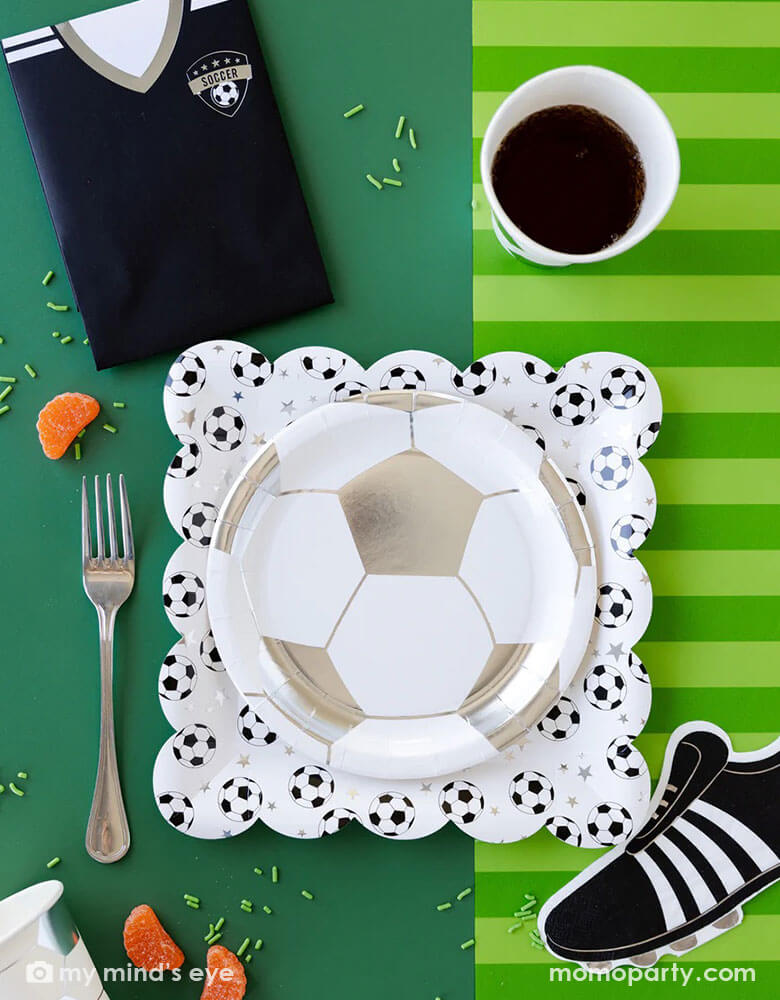 A fun soccer themed party table featuring Momopartys's soccer party collection including silver soccer shaped plates and party cups, cleat shaped napkins, soccer jersey shaped treat bags and soccer table runner by My Mind's Eye. With some snacks, green sprinkles around, this makes a great inspo for a fun kid's soccer themed birthday party or a championship celebration!