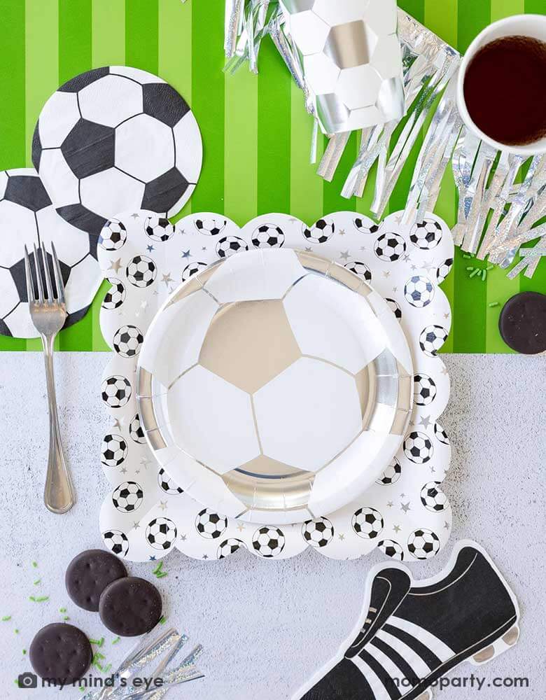 A fun soccer themed party table featuring Momopartys's soccer party collection including silver soccer shaped plates and party cups, soccer shaped napkins, cleat shaped napkins and soccer table runner by My Mind's Eye. With some snacks, silver streamers, green confetti and silver utensil around, this makes a great inspo for a fun kid's soccer themed birthday party or a championship celebration! 