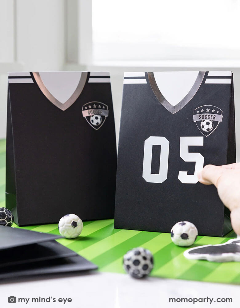 A soccer themed party table features Momopartys's black soccer jersey shaped treat bags with number stickers by My Mind's Eye. On the table there's a green striped soccer table runner and some soccer ball shaped chocolate balls, making it perfect for kid's soccer themed birthday party or championship celebration bash!