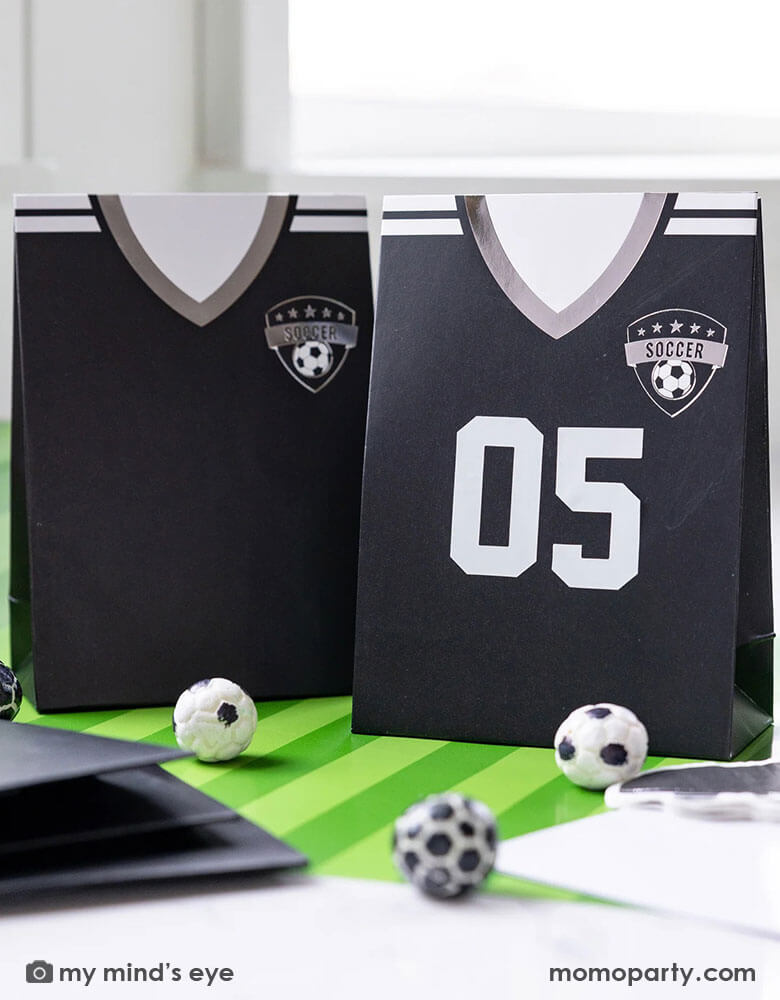 A soccer themed party table features Momopartys's black soccer jersey shaped treat bags with number stickers by My Mind's Eye. On the table there's a green striped soccer table runner and some soccer ball shaped chocolate balls, making it perfect for kid's soccer themed birthday party or championship celebration bash!