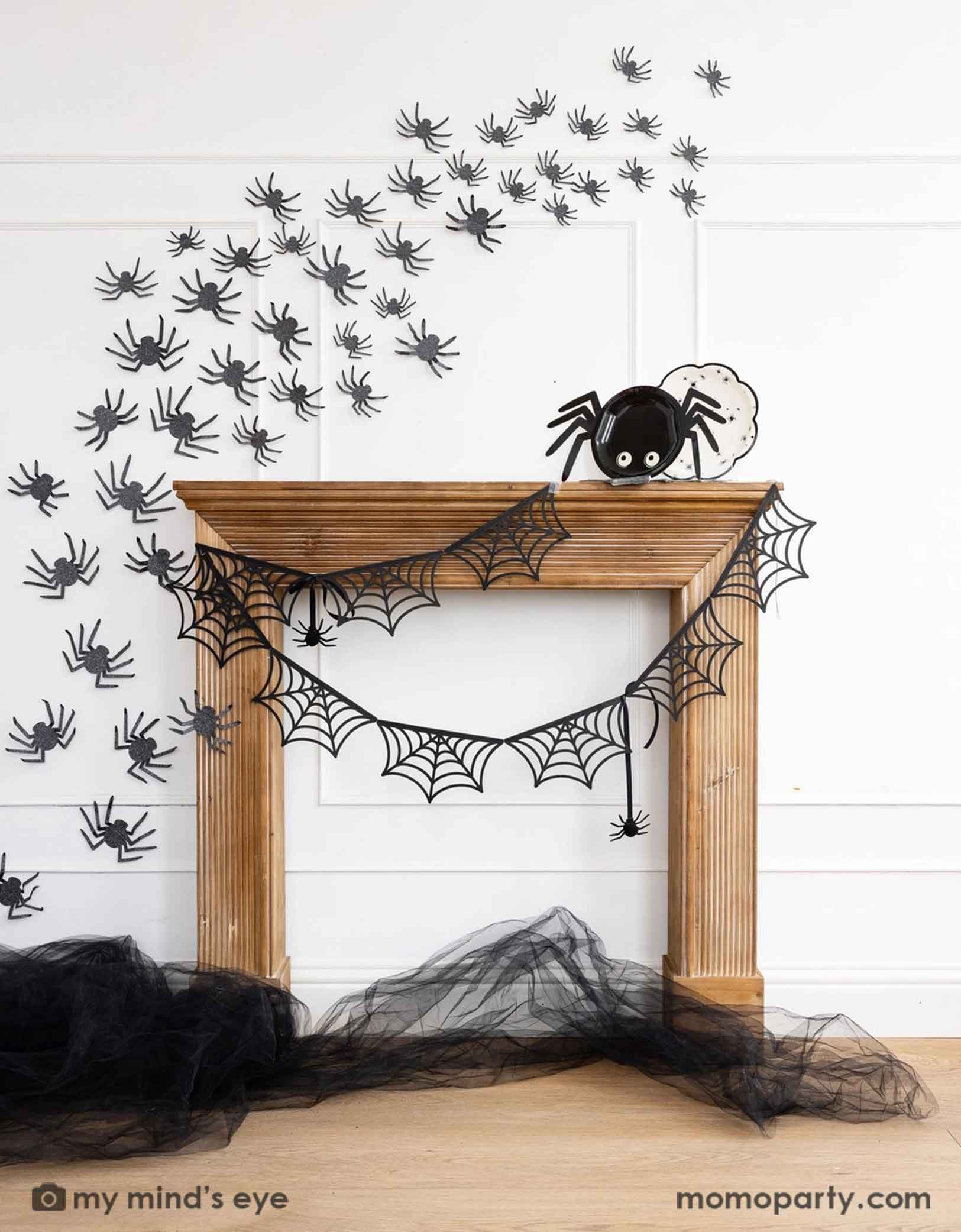 A wooden mantel against a white wall decorated with Halloween decorations from Momo  Party including the spider web banner, black spider decorations on the wall and the 14" x 7" spider shaped plate and a round plate with spider designs on it. On the floor there's some black tulle on the floor, making this a kid's friendly cute Halloween decorating idea for the spooky season. 