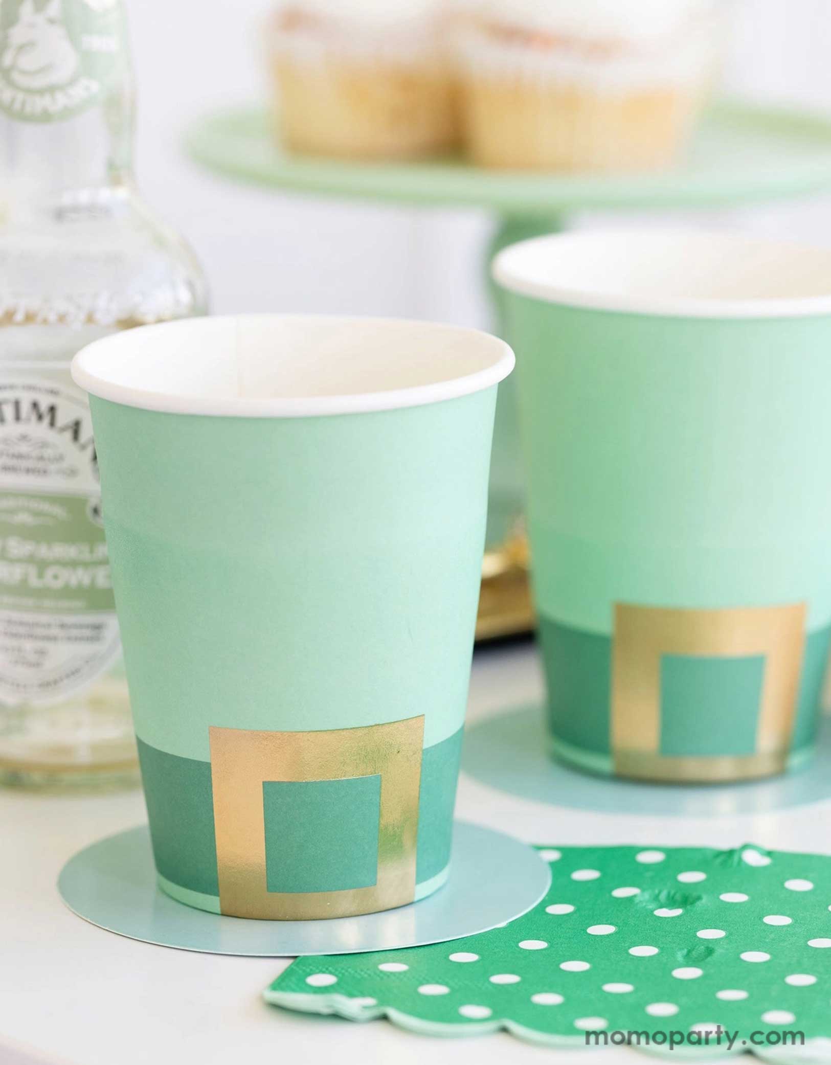 A cheerful St. Patrick's Day party table featuring Momopartys's St. Patrick's Day themed party supplies including the Leprechaun hat party cups and green polka dot napkins. In the back there's a green cake stand with cupcakes on a gold serving tray. With drink bottles next to them making this a fun and festive St. Patrick's Day celebration.