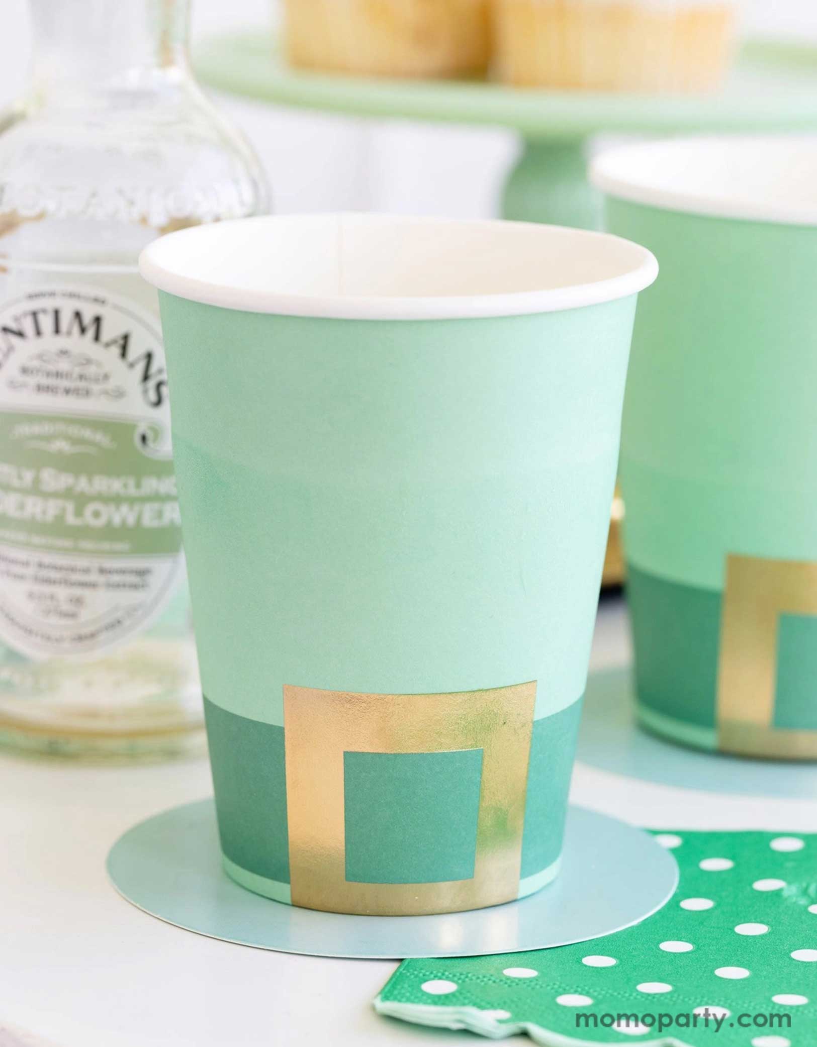 A cheerful St. Patrick's Day party table featuring Momopartys's St. Patrick's Day themed party supplies including the Leprechaun hat party cups and green polka dot napkins. In the back there's a green cake stand with cupcakes on a gold serving tray. With drink bottles next to them making this a fun and festive St. Patrick's Day celebration.