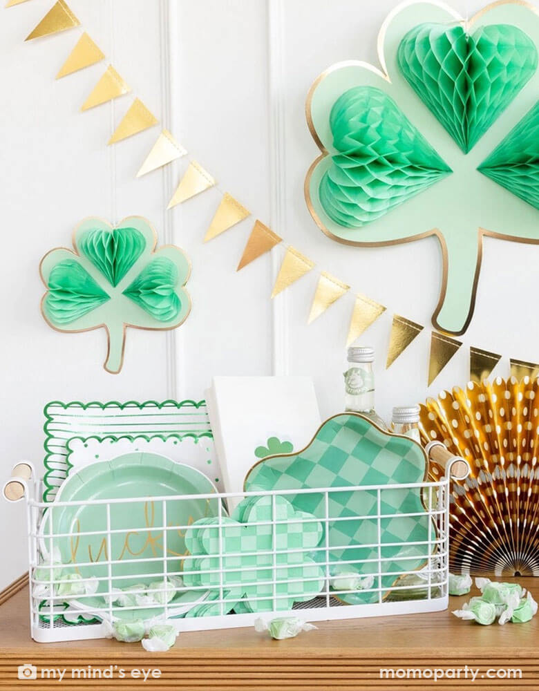 A corner of a house decorated with Momopartys's shamrock honeycomb decorations and a gold foil flag banner hung on the wall. On the cabinet counter, there's a gold foil paper fan and a basket filled with Momopartys's St. Patrick's Day party tableware including scalloped paper plates, lucky paper plate set, checkered shamrock shaped napkins and plates next to some green taffy candies, makes this a perfect info for a festive St. Patrick's Day celebration. 