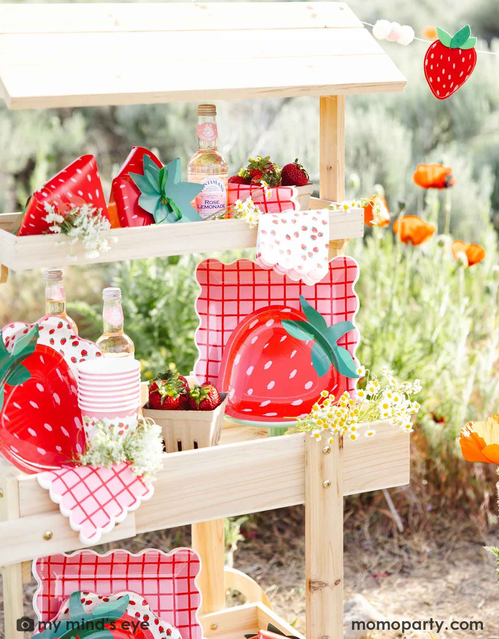 A wooden dessert cart in an outdoor field filled with strawberry-themed tablewares and decorations from Momopartys including strawberry felt banner, strawberry shaped plates, strawberry scattered guest napkins, party cups and treat boxes. Along with bottles, flowers, and strawberries in small baskets, this farmers market-like set up is adorable for kid's "Berry First Birthday" strawberry themed 1st birthday party celebration.