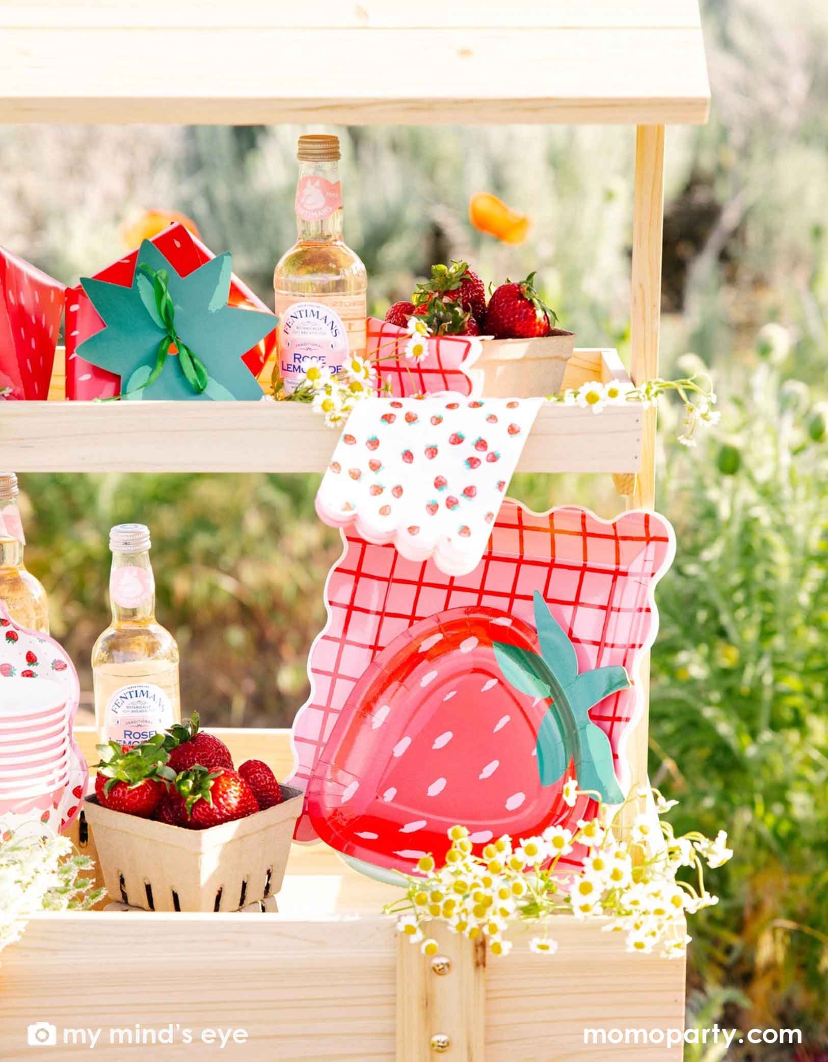 A wooden display cart in an outdoor field filled with strawberry-themed tablewares and decorations from Momopartys including strawberry shaped plates, strawberry scattered guest napkins, party cups and treat boxes. Along with bottles, flowers, and strawberries in small baskets, this farmers market-like set up is adorable for kid's "Berry First Birthday" strawberry themed 1st birthday party celebration.