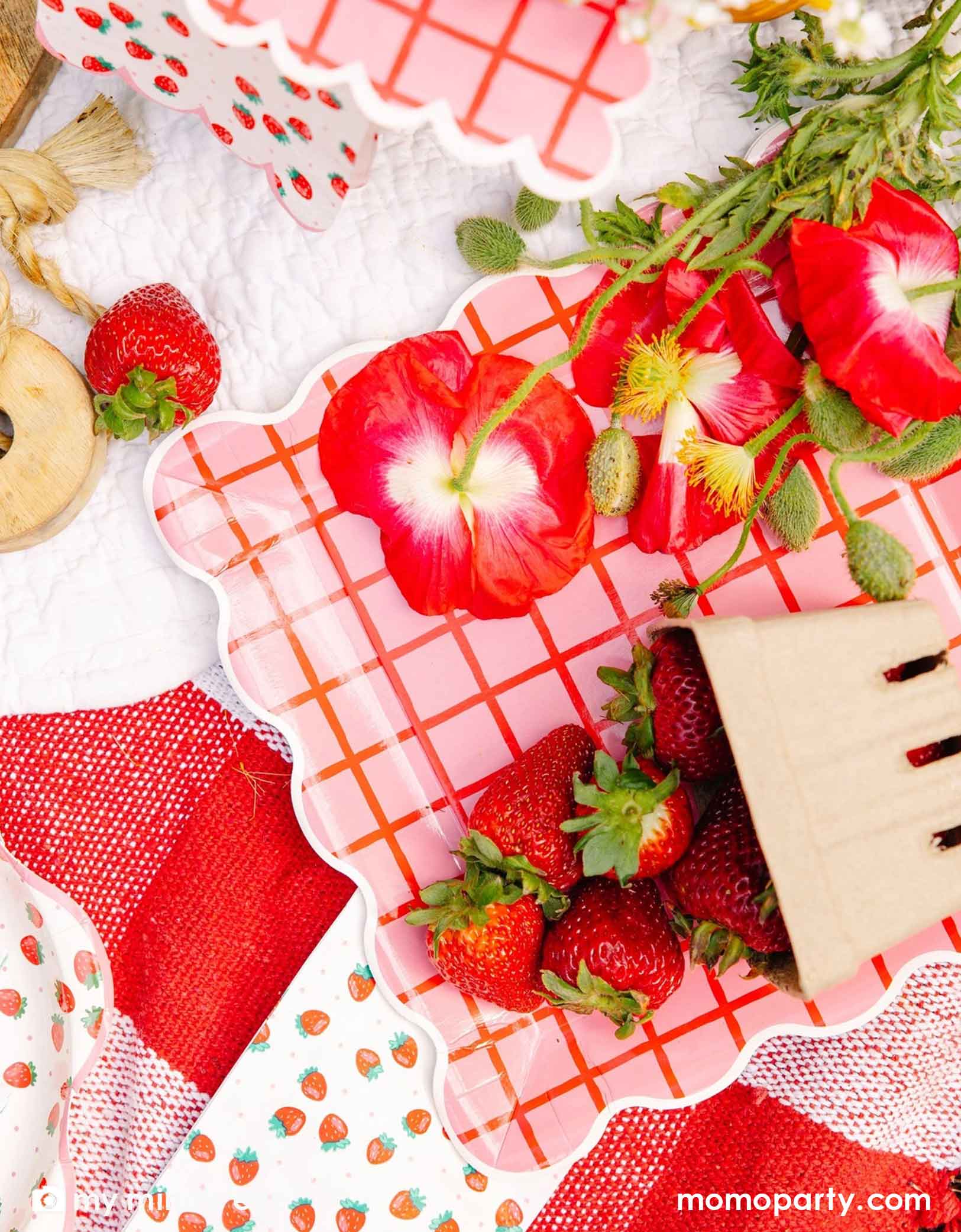 An overhead shot of a strawberry themed tablescape featuring Momopartys's strawberry tablewares including the 10" red and pink grid paper plates, strawberry patterned napkins and plates. Along with red flowers and strawberries scattered around on a white surface.