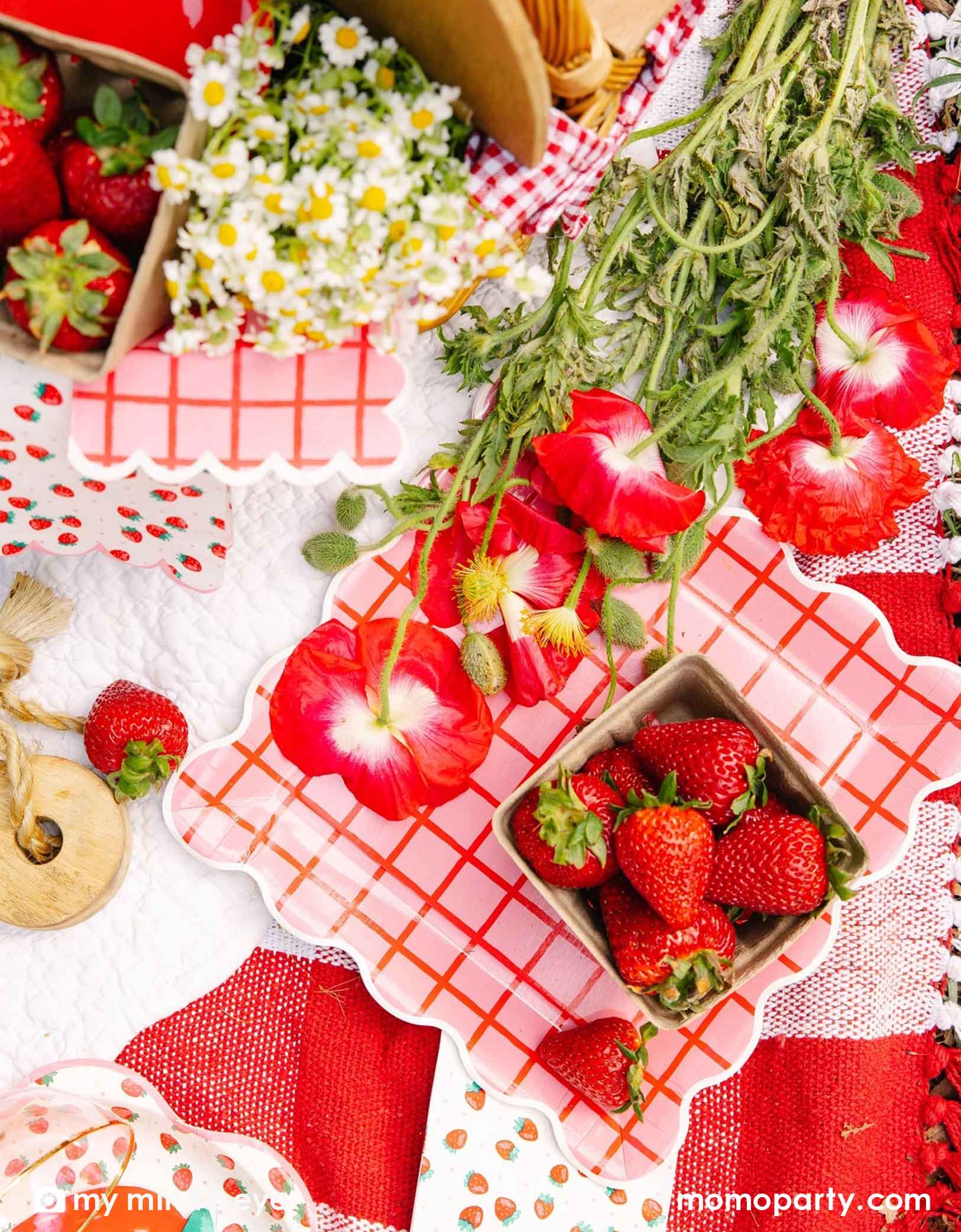 An overhead shot of a strawberry themed tablescape featuring Momopartys's strawberry tablewares including the 10" red and pink grid paper plates, 5"x5" napkins, strawberry patterned napkins and plates. Along with red flowers and strawberries scattered around on a white surface.