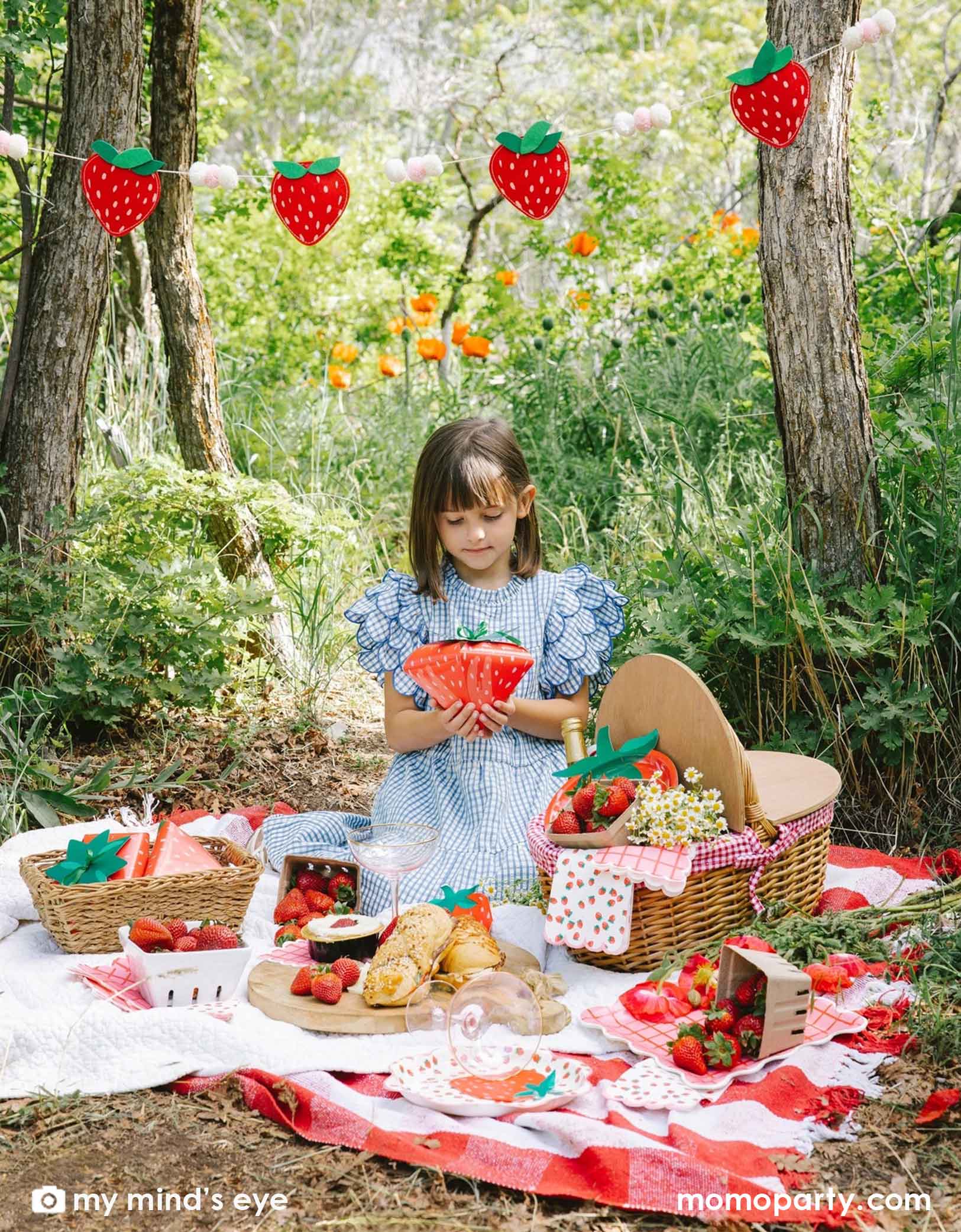 A girl in a blue gingham dress sitting at a picnic blanket in a forest with a decorated area and strawberry decorations and party tablewares from Momopartys including strawberry felt party banner, strawberry plates, napkins, paper cups and strawberry shaped treat boxes. A whimsical and adorable setting for a special celebration. 