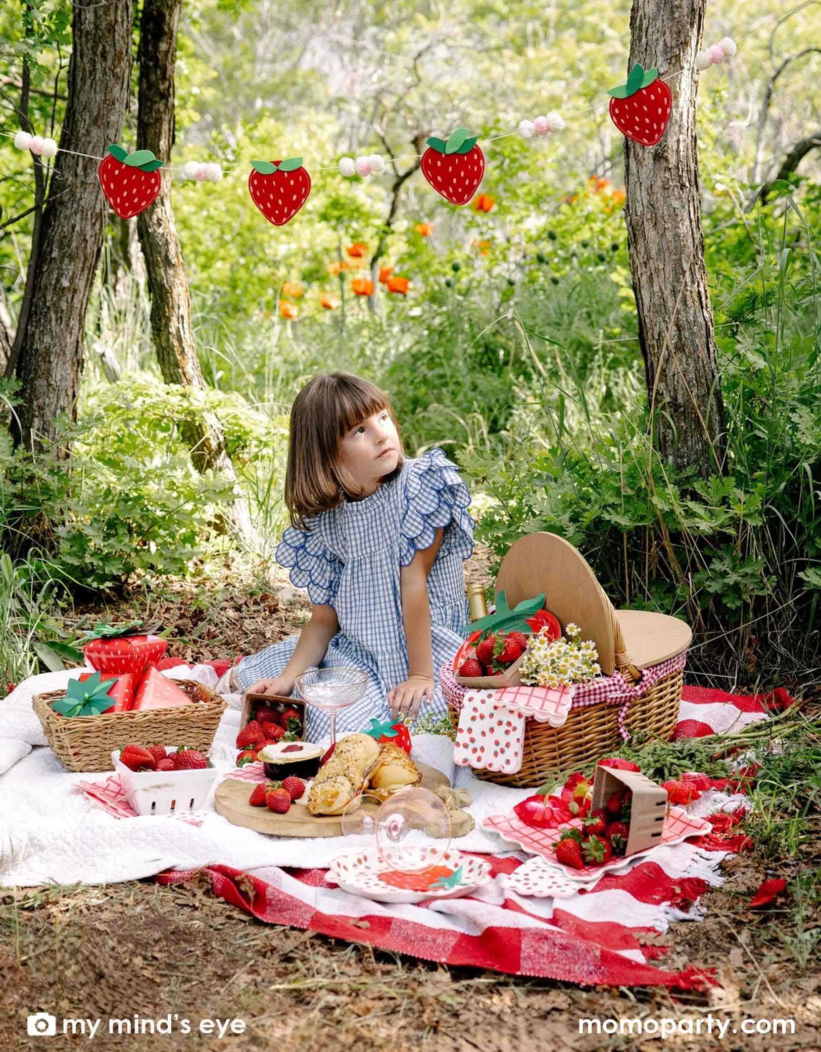 A girl in a blue gingham dress sitting at a picnic blanket in a forest with a decorated area and strawberry decorations and party tablewares from Momopartys including strawberry felt party banner, strawberry plates, napkins, paper cups and strawberry shaped treat boxes. A whimsical and adorable setting for a special celebration. 