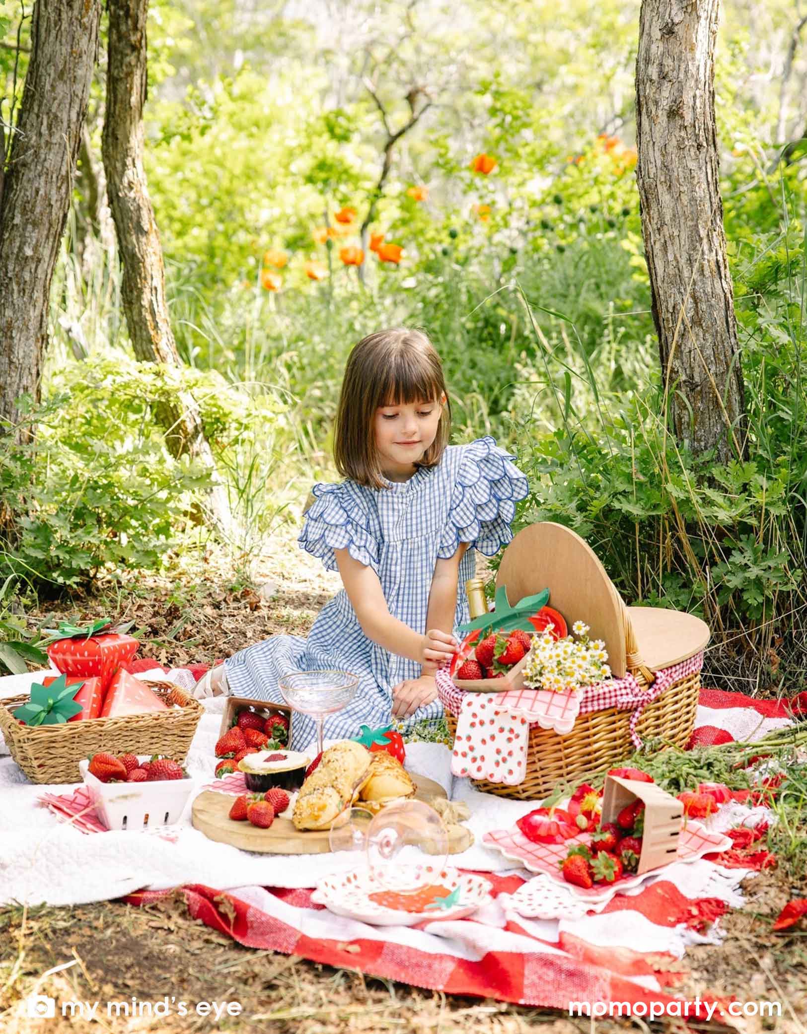 A girl in a blue gingham dress sitting at a picnic blanket with a picnic basket in a forest with a decorated area and strawberry decorations and party tablewares from Momopartys including 10" strawberry plates, napkins, paper cups and strawberry shaped treat boxes. A whimsical and adorable setting for a special celebration. 
