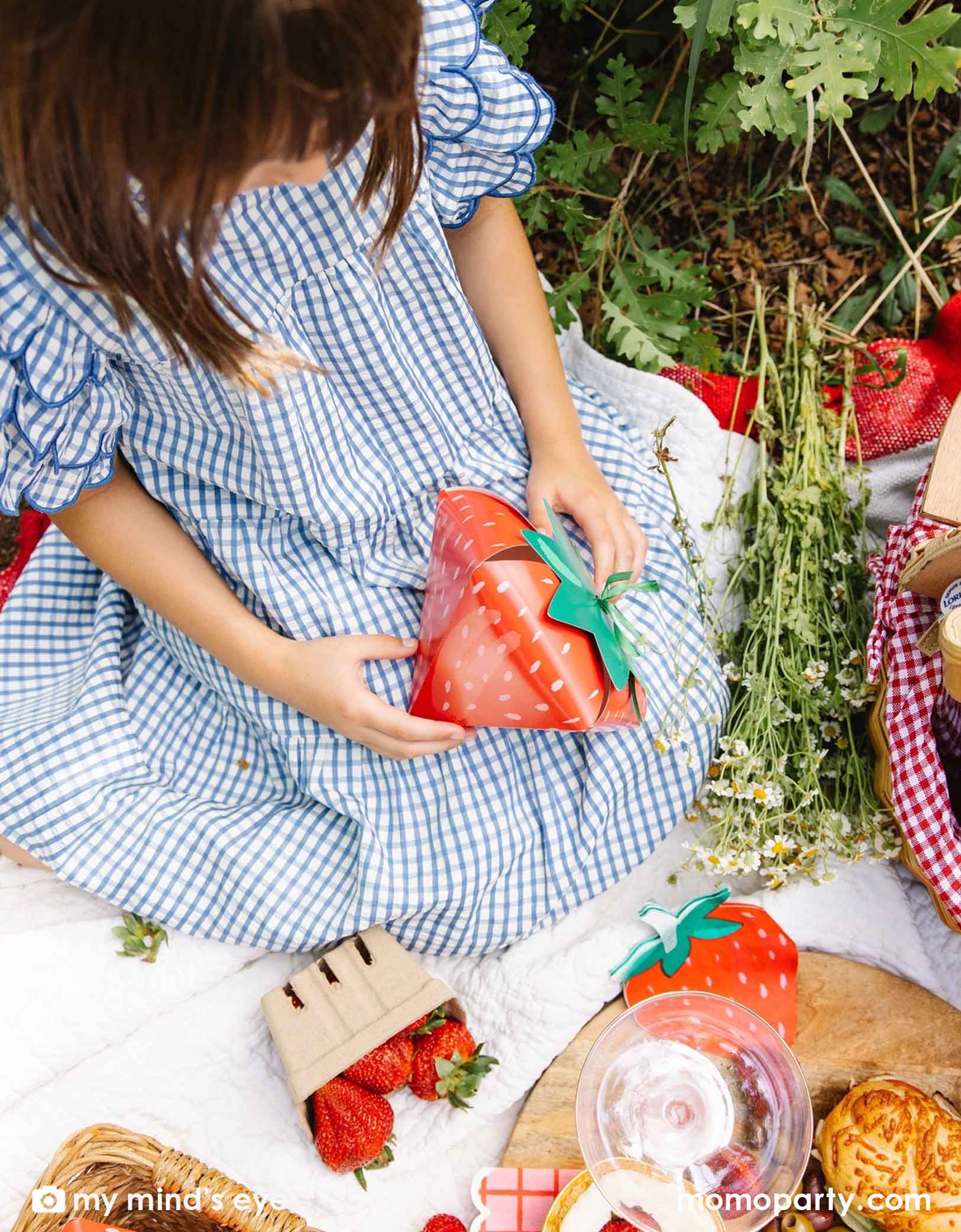 A girl in a blue gingham dress holding a strawberry-shaped treat box by Momopartys sitting on a picnic blanket with various strawberry themed party tablewares including strawberry shaped napkins, plates and party cups.