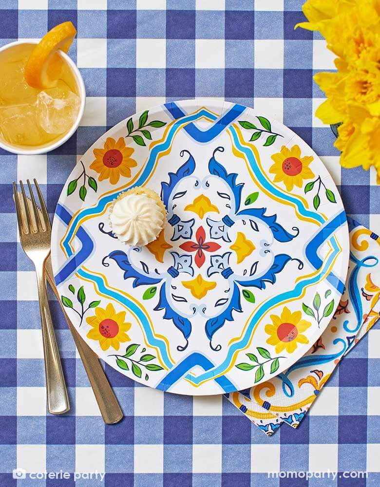 A flatlay view of a summer party table features Momopartys's navy gingham tablecover by Coterie and summer themed paper plates and napkins on it, next to the tableware there are some yellow flowers and a drink topped with an orange slice. Making this a perfect inspo for a stylish summer party tablescape. 