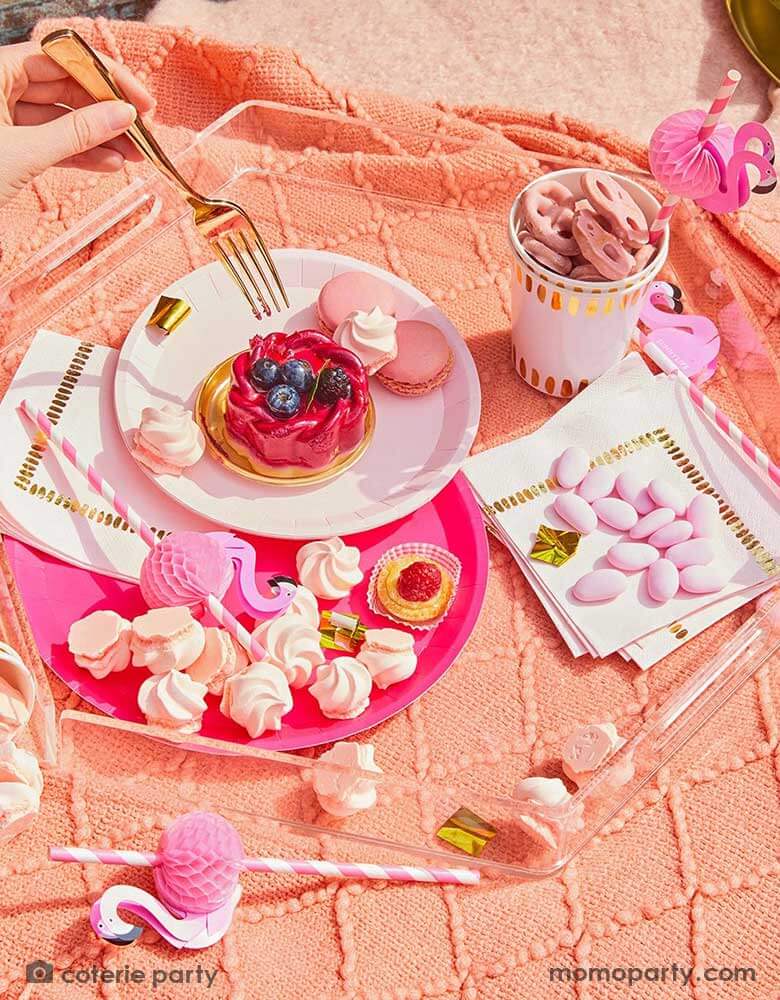 A summer picnic party under the sunset featuring Momopartys's summer party supplies including hot pink dinner plates, pale pink side plates, modern white party cups and napkins with gold foil accents. On the plates are some summer treats, fruit tarts, cookies and meringues, along with flamingo honeycomb paper straws - all on a coral colored picnic blanket. Making this a perfect inspo for a modern yet festive summer gathering. 