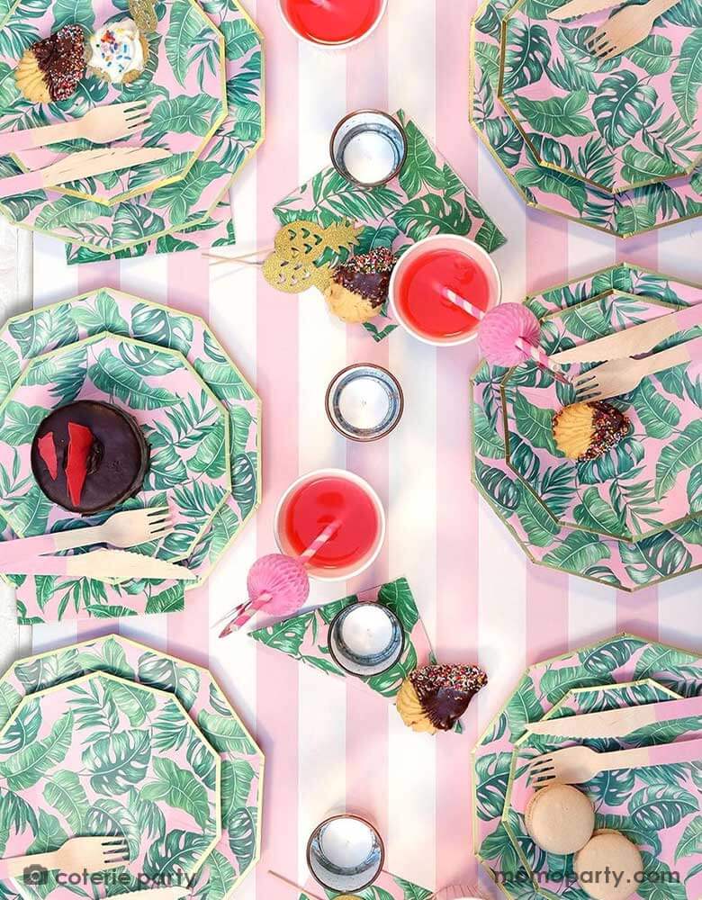 A modern summer tropical themed party tablescape features Momopartys's tropical party themed party supplies including palm leaf plates, napkins, pink wooden utensil set, pineapple shaped treat toppers and flamingo honeycomb paper straws in the party cups filled with pink drinks All these are on a pink striped table runner, making this a festive yet modern look for a summer party!