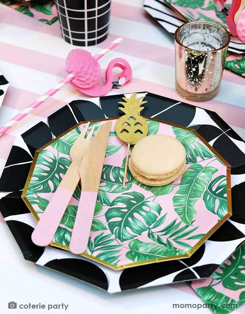 A closeup shot of a modern summer tropical themed party placeset features Momopartys's tropical party themed party supplies including palm leaf plates, napkins, pink wooden utensil set and flamingo honeycomb paper straws in the party cups. All these are on a pink striped table runner, making this a festive yet modern look for a summer party!