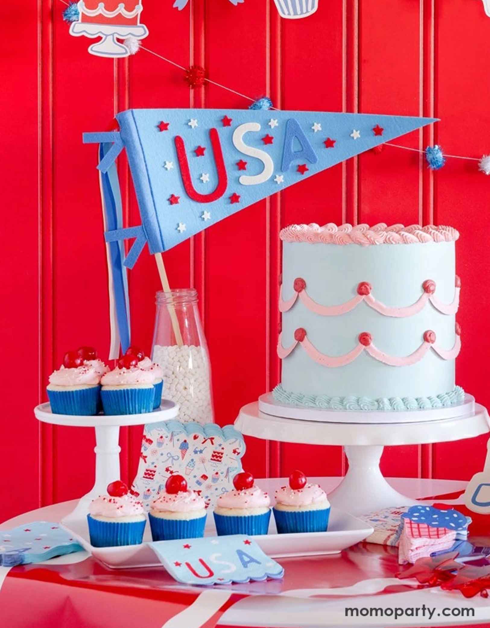 A festive patriotic party set up featuring various Fourth of July themed party decors from Momopartys including party banner with summer themed party pennants including popsicles, ice cream, cupcake, cakes, and star shaped sunglasses in red, blue and white colors. On the table there's a vintage pastel blue buttercream cake along with some cupcakes with cherries on top, along with a USA felt pennant and patriotic party tableware including party plates, napkins and a table runner, for a fun summer party.