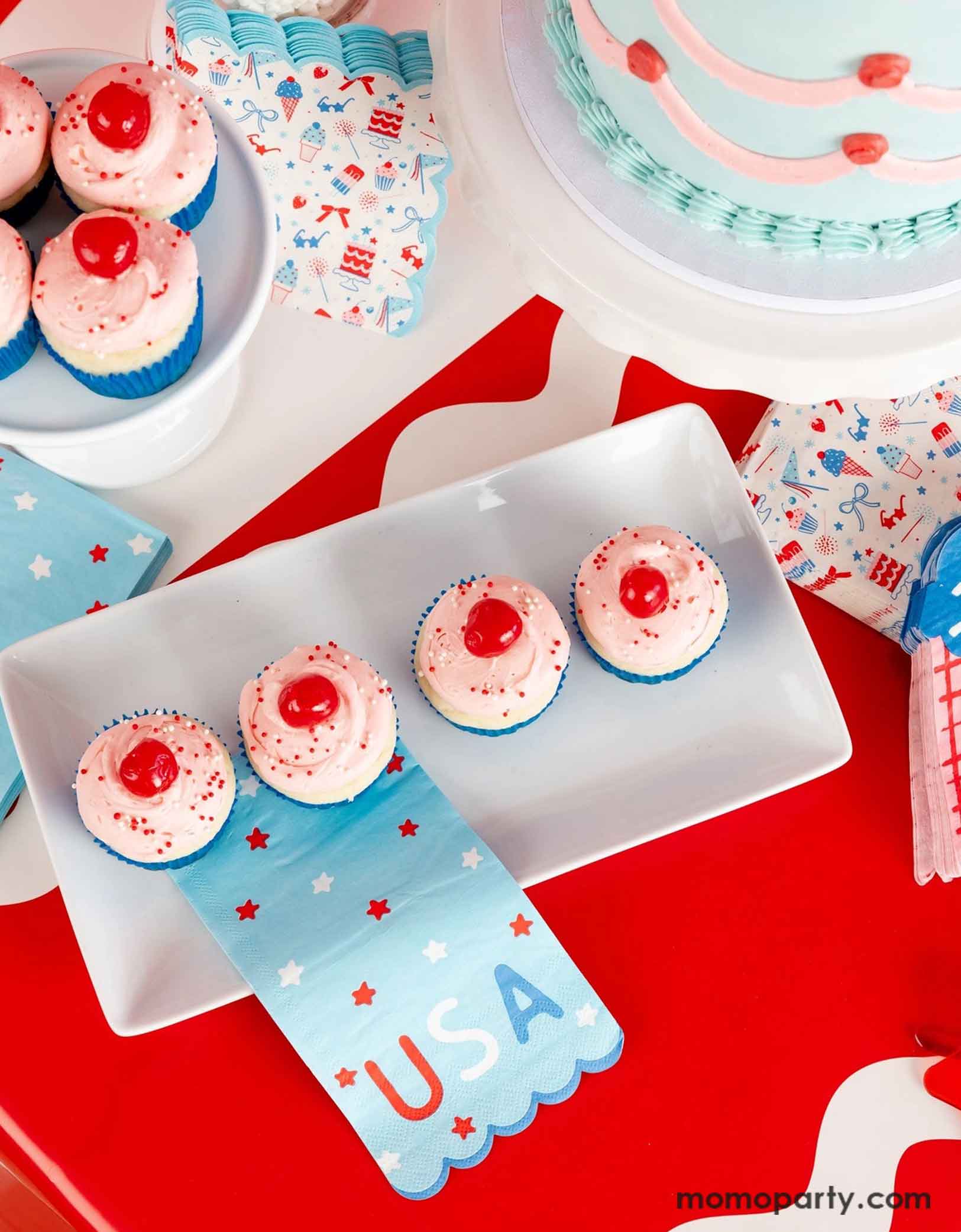 An overhead shot of a festive patriotic themed or a Fourth of July summer party featuring Momopartys's USA inspired party supplies and colorful treats and desserts including cupcakes with cherries on them and a vintage style buttercream cake. Around the treats are some Independence Day inspired party napkins in a modern style on a red table runner. 